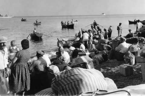 L'immagine in bianco e nero AP mostra le famiglie con borse raccolte sulla spiaggia. Alcuni vagano per le distanze in piccole barche a remi di legno